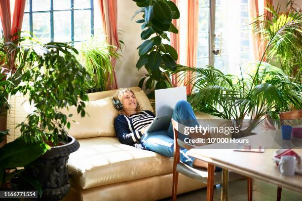 mature woman smiling and relaxing on sofa with headphones - comfortable retirement stock pictures, royalty-free photos & images