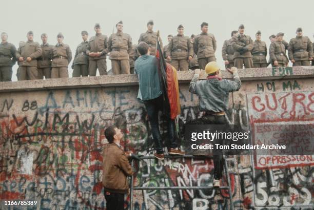 East German border guards stand in line on the Berlin Wall as as West Berliners with a German flag scale the wall on the morning of 10th November...