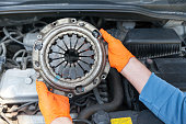 Auto mechanic wearing protective work gloves holding used clutch pressure plate above a car engine
