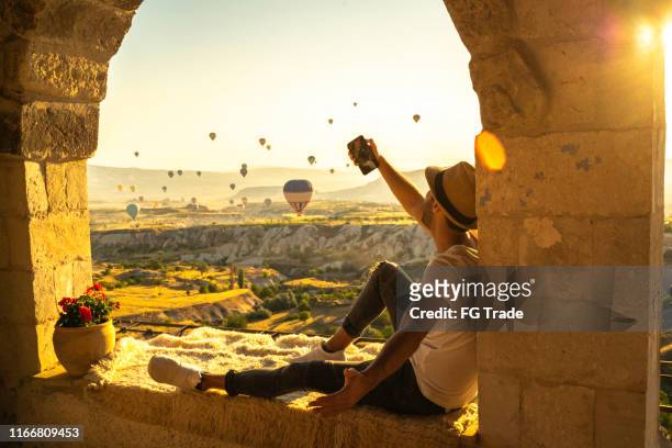 side view of a young man taking a selfie sitting and looking at view during ballooning festival - göreme stock pictures, royalty-free photos & images