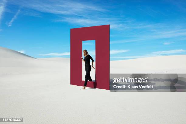 young fashion model walking through red door frame at desert against sky - tür stock-fotos und bilder