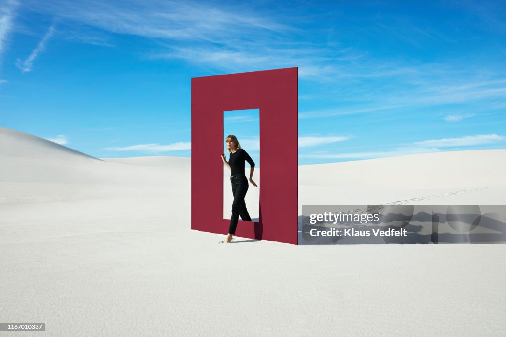 Young fashion model walking through red door frame at desert against sky