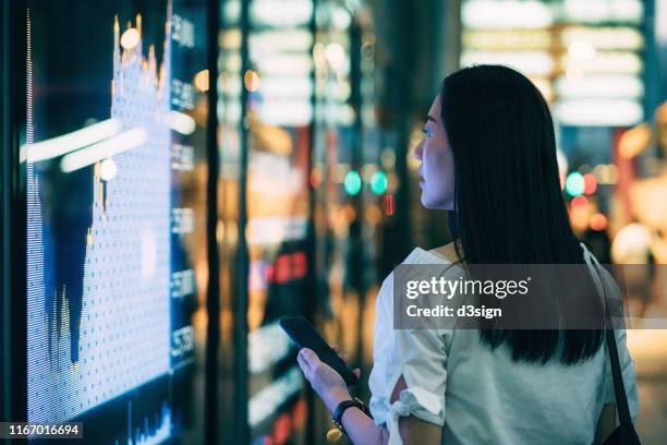 young businesswoman checking financial trading data on smartphone by the stock exchange market display screen board in downtown financial district - china economy stockfoto's en -beelden