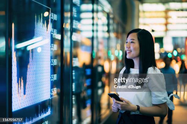 confidence young asian businesswoman checking financial trading data on smartphone by the stock exchange market display screen board in downtown financial district - financial market photos et images de collection