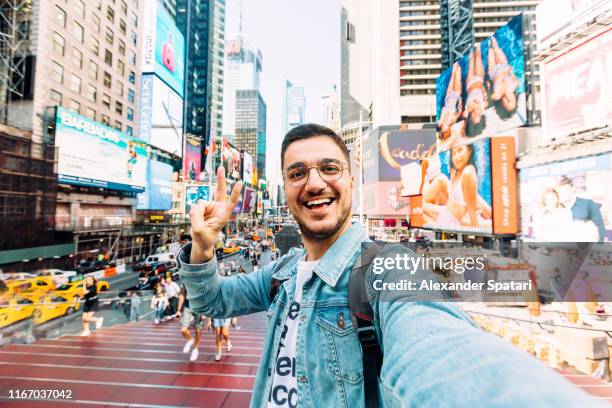 young happy man taking selfie and showing peace gesture at times square, new york city, usa - call us photos et images de collection