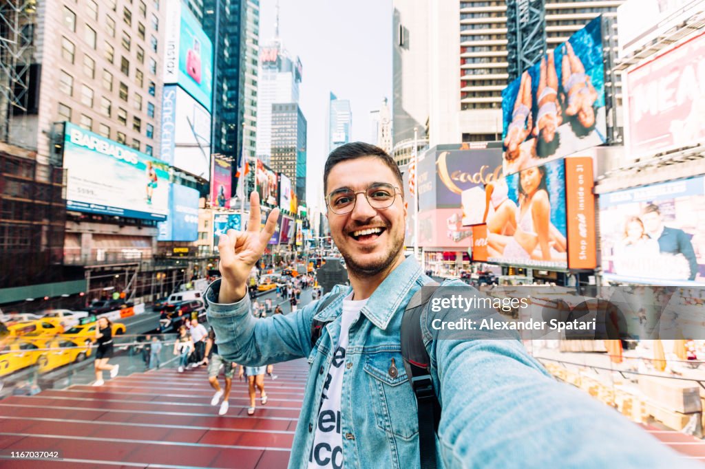 Young happy man taking selfie and showing peace gesture at Times Square, New York City, USA