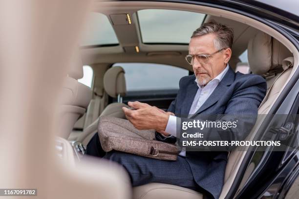overworked businessman sitting on back seat in car and using mobile phone - laptoptasche stock-fotos und bilder