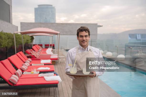 männlicher kellner, der eine bestellung von getränken hält, während er in die kamera schaut und lächelnd in einem luxushotel neben dem pool auf dem dach steht - rooftop pool stock-fotos und bilder