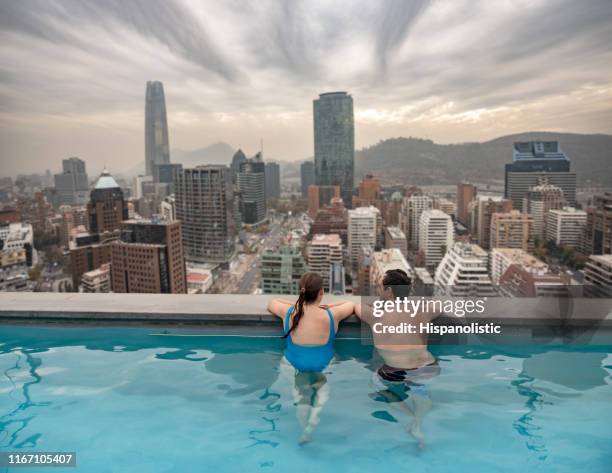 lateinamerikanisches paar genießt den pool am rande des pools mit blick auf eine schöne stadtblick vom dach - rooftop pool stock-fotos und bilder