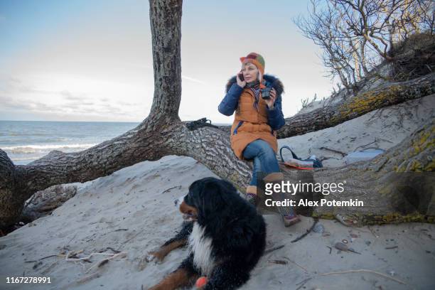 die 55-jährige reife spricht per smartphone und trinkt einen heißen tee und ruht mit ihrem zennenhund hund an der ostseeküste. - 55 years old stock-fotos und bilder