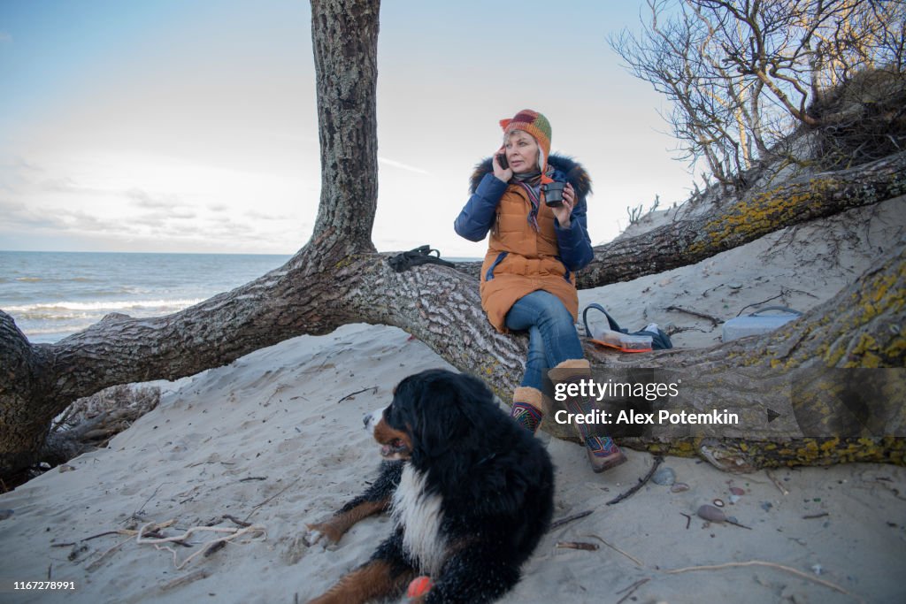 La mujer madura de 55 años hablando a través de un teléfono inteligente y bebiendo un té caliente, descansando en la orilla del mar Báltico con su perro Zennenhund.