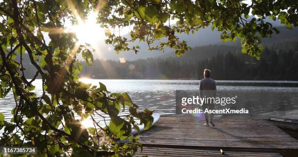 man walks onto dock over lake and watches sunrise over mountains and forest - tranquility stock pictures, royalty-free photos & images