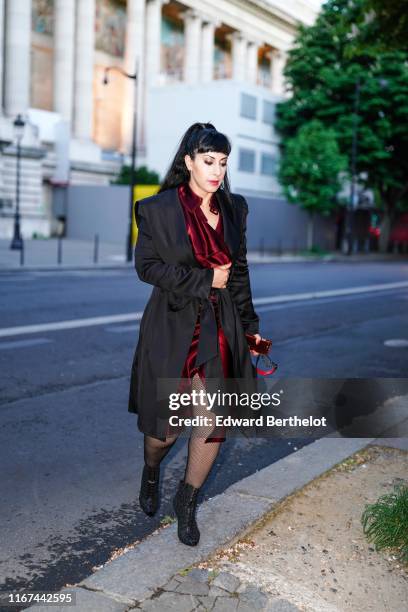 Guest wears a shiny burgundy satin front split dress, a lustrous oversize black coat, black fishnet tights, black openwork peep-toed heeled ankle...