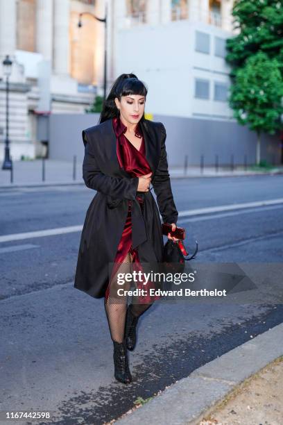 Guest wears a shiny burgundy satin front split dress, a lustrous oversize black coat, black fishnet tights, black openwork peep-toed heeled ankle...