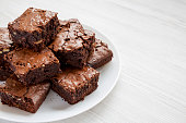 Homemade chocolate brownies on a white plate on a white wooden background, side view. Copy space.