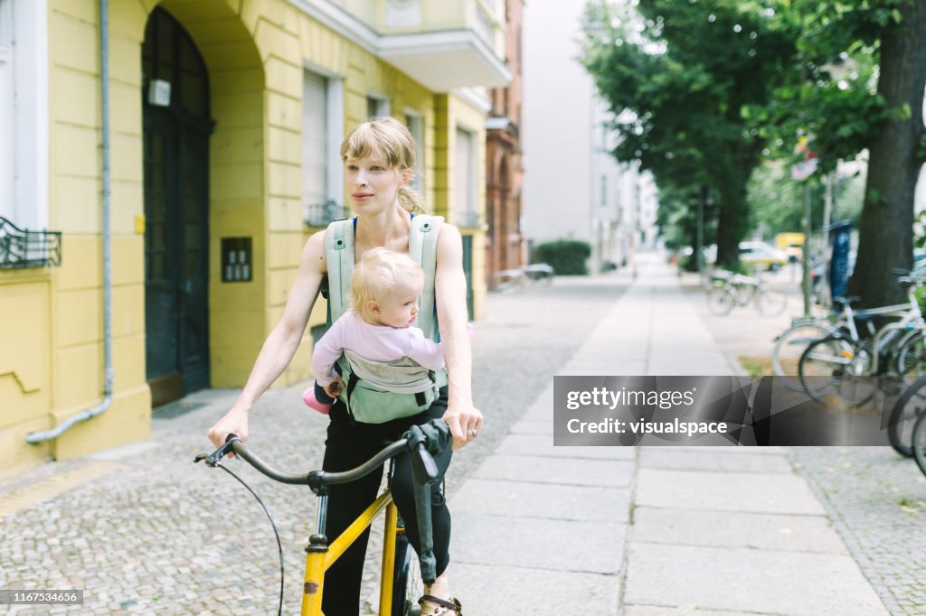 Jeune mère avec le bébé dans le porteur conduisant le vélo par la ville