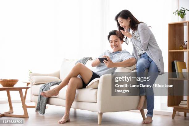 happy young chinese couple using smartphone in living room - happy couple using cellphone stockfoto's en -beelden