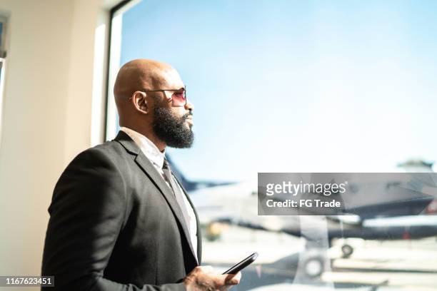 empresario de pie junto a la ventana y contemplando, sosteniendo el teléfono inteligente en el aeropuerto - best sunglasses for bald men fotografías e imágenes de stock