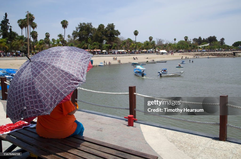 Woman with purple patterned umbrella on a park bench overlooking beach and boats on Lake Chapala; Chapala, Jalisco, Mexico
