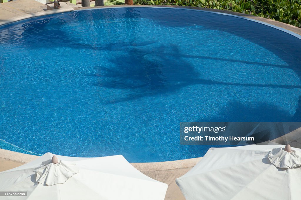 Tops of two white umbrellas in foreground, round swimming pool with palm tree shadows beyond; Costalegre, Jalisco, Mexico