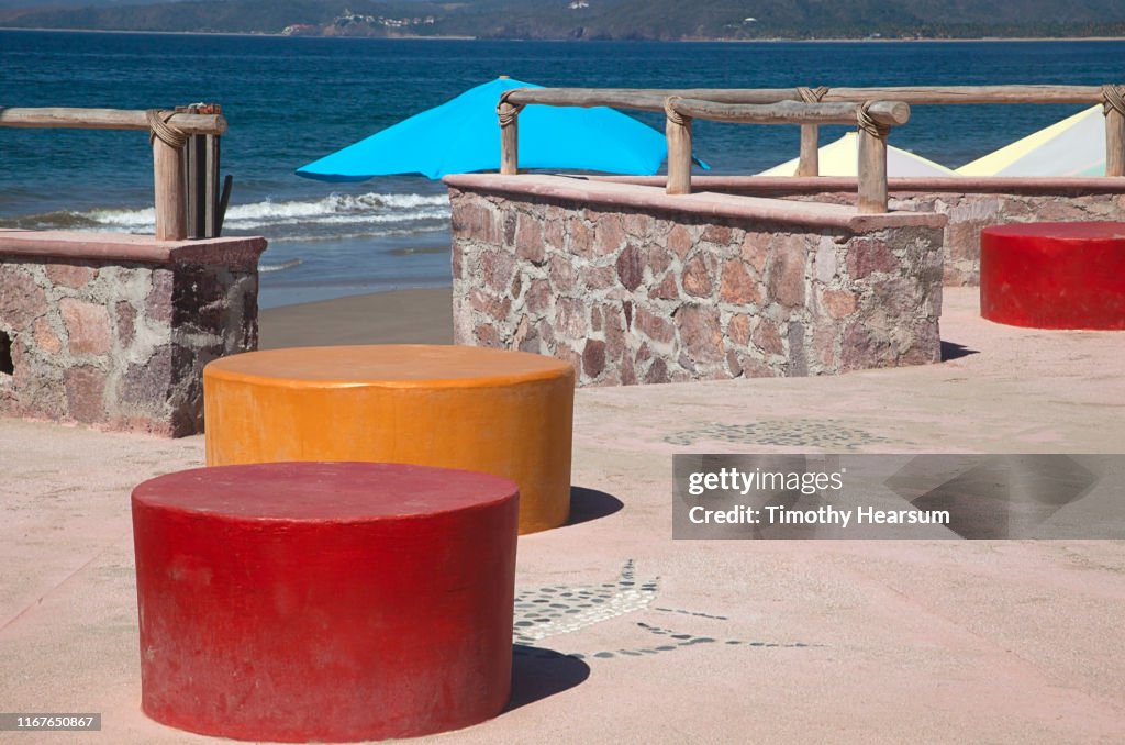 Red and Orange round benches on a patio in foreground; beach umbrellas and ocean beyond; Tenacatita Bay, Costalegre, Jalisco, Mexico