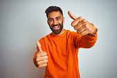 Young indian man wearing orange sweater over isolated white background approving doing positive gesture with hand, thumbs up smiling and happy for success. Winner gesture.