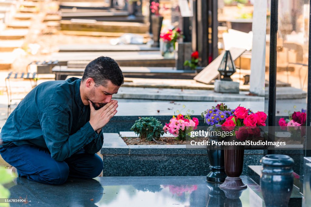 Emotionally Stressed Man in Black Kneeling in Front of Grave of his Family Member