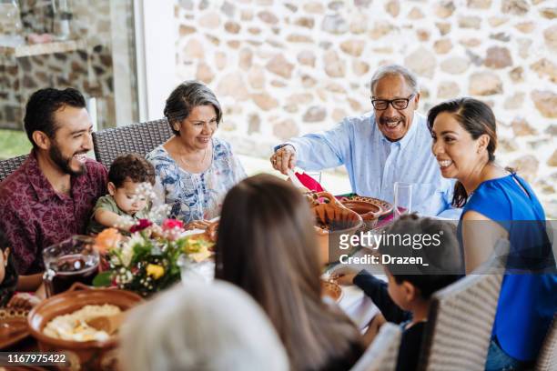latin senior man serving the food to his family at dinner table - multi generation family stock pictures, royalty-free photos & images