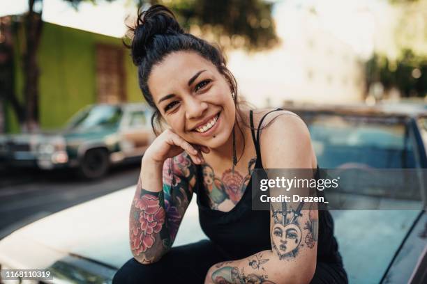 tattooed latina smiling outdoors and sitting on car hood - tattoo woman stock pictures, royalty-free photos & images