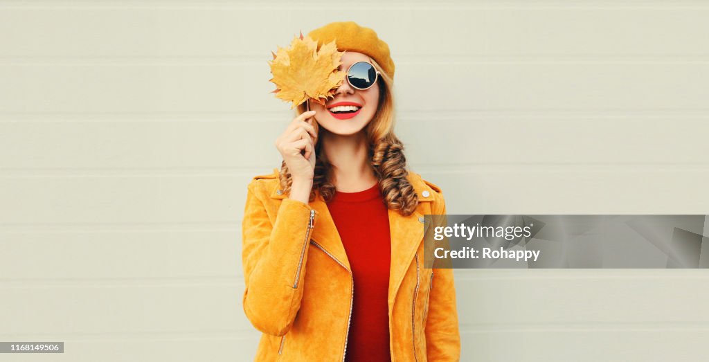 Autumn mood! happy smiling woman holding in her hands yellow maple leaves hiding her eye over gray wall background
