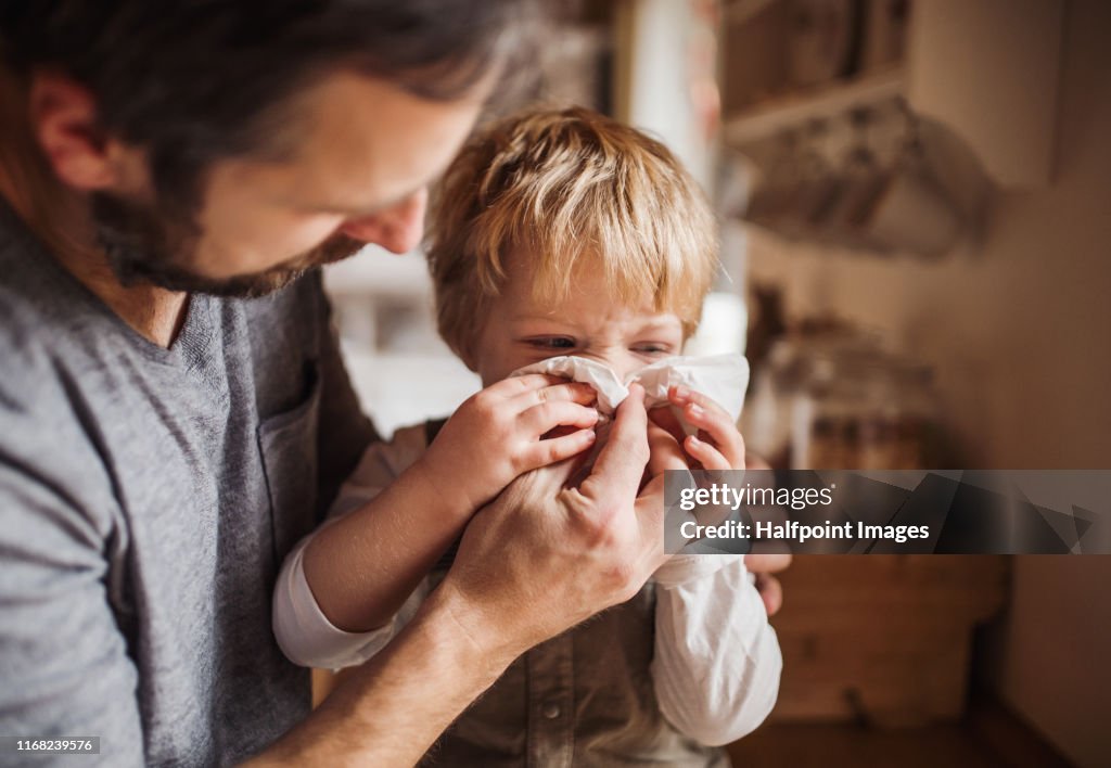 A father on paternity leave looking after small son indoors, blowing his nose.