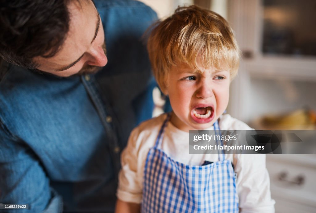 A father on paternity leave talking to crying small son indoors.