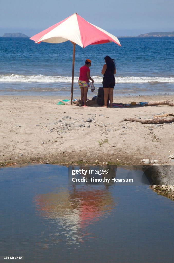 Two women standing under red and white umbrella on a beach on Tenacatita Bay; Costalegre, Jalisco, Mexico