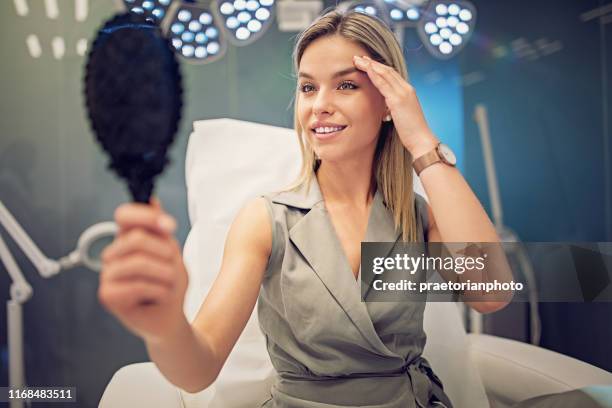 la jeune femme regarde son visage dans le miroir après la procédure de traitement de beauté - chirurgie esthétique photos et images de collection