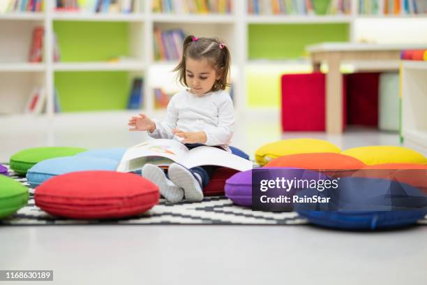 ragazza carina sta leggendo un libro seduto nella biblioteca del vivaio - ludoteca foto e immagini stock
