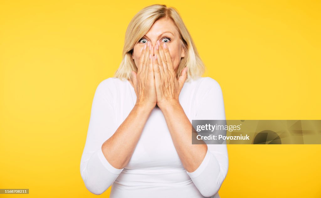 Don't talk! Blonde Senior woman covers her mouth with help of hands and looks on camera with shock in eyes.