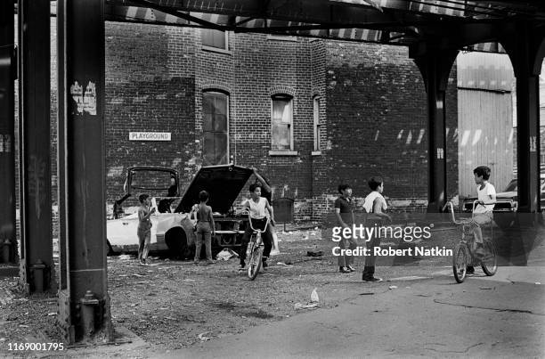 Near a sign that reads 'Playground,' two kids ride bicycles while others gather around an abandoned convertible in a lot under the elevated train...
