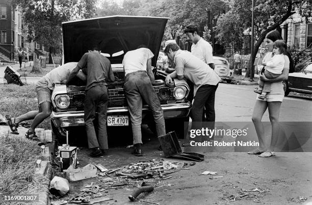 Young woman with a child watches as a group of men under the open hood of a car as they work on its engine, Chicago, Illinois, 1970s. A can of...