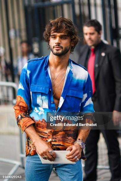 Marlon Teixeira wears necklaces, a landscape photo print shirt, rings, bracelets, blue denim pants, outside Balmain, during Paris Fashion Week -...