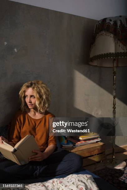 blond student sitting on bed with a book looking at distance - pensive man stock-fotos und bilder