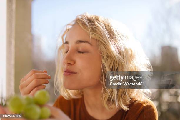 portrait of blond young woman eating green grapes - positive emotionen stock-fotos und bilder