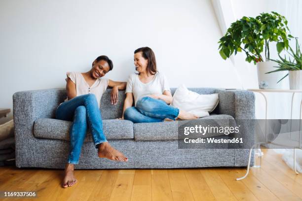 two happy relaxed women sitting on couch at home - couple sitting stock pictures, royalty-free photos & images