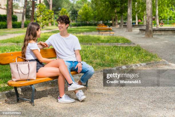 young couple sitting on a bench and talking to each other in a park - sitting bench stock pictures, royalty-free photos & images