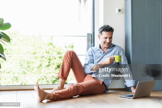 mature man relaxing at home with a cup of coffee, using laptop - auf dem boden sitzen stock-fotos und bilder