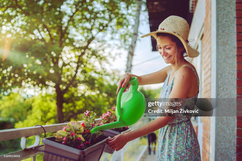 Joven hermosa mujer regando las flores de las plantas en su balcón de la casa usando cubo con agua usando vestido de verano y sombrero en el día de la noche de otoño de primavera