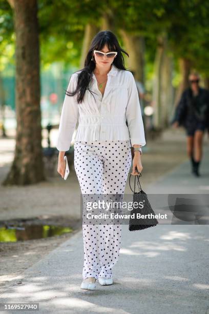 Guest wears earrings, white sunglasses, a white pleated shirt, white pants with black polka dots, a black Prada net bag, white shoes with separated...