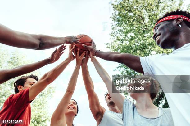 low-winkel-ansicht von lächelnden jungen männlichen und weiblichen freunden, die basketball auf dem platz gegen den himmel halten - basketballmannschaft stock-fotos und bilder