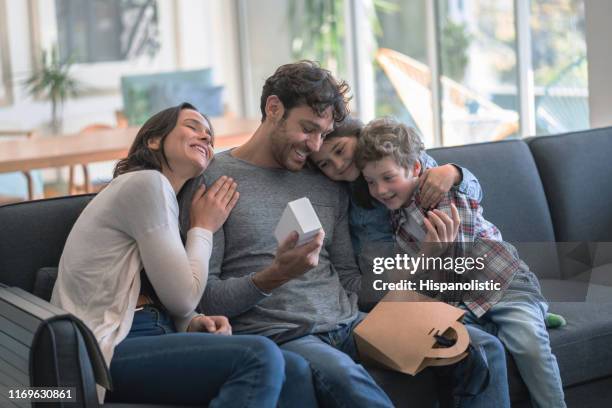 amorevole madre e figli che celebrano la festa del padre abbracciando papà mentre opne un regalo tutto sorridente - festa del papà foto e immagini stock