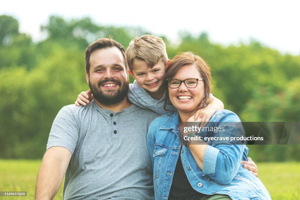 Millennial Vater und Mutter mit autistischen Kind in öffentlichen üppigen grünen Park sitzt auf dem Gras lächelnd Pose für Familienporträt