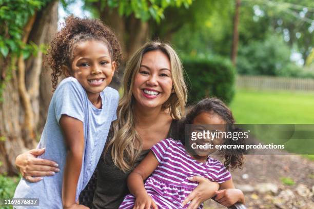 asian chinese mother with two daughters of mixed chinese and african american ethnicity in a green lush back yard setting posing for portraits smiling and being silly - foster stock pictures, royalty-free photos & images